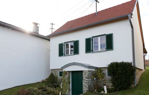 Ein weißes Haus mit rotem Dach und grünen Fensterläden ist von Grün umgeben. Unten befinden sich die Fenster ein grüner Eingang.