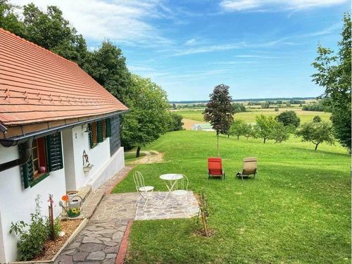 Ein Landhaus mit rotem Dach und grünen Fensterläden steht in einem großen Feld. Ein Patio hat einen Tisch und zwei Stühle. In der Ferne eine Baumreihe und ein grünes Feld.