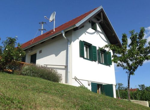 Ein weißes Haus mit rotem Dach und grünen Fensterläden steht auf einem Grashügel. Es hat eine Satellitenschüssel und einen Schornstein.