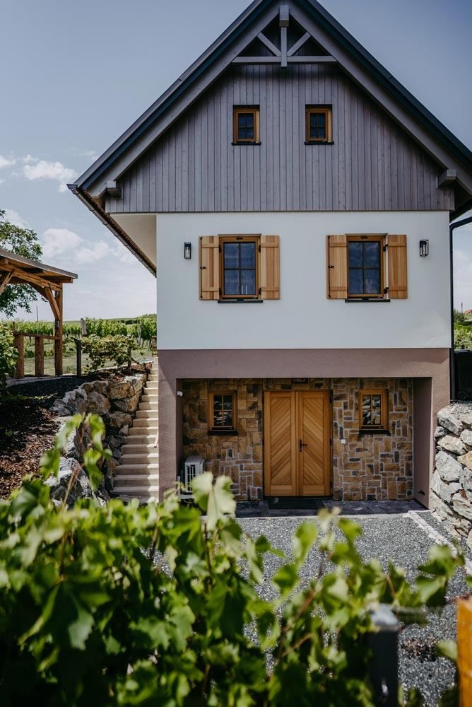 Ein zweistöckiges Haus mit einem grauen Holzoberteil und einem Steinziegelunterteil. Es hat Holztüren, Glasfenster und Holzläden. Es gibt eine Steinmauer mit Treppen und einen hölzernen Pavillon.