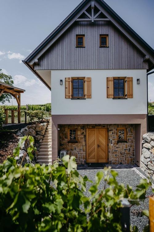 Ein zweistöckiges Haus mit einem grauen Holzoberteil und einem Steinziegelunterteil. Es hat Holztüren, Glasfenster und Holzläden. Es gibt eine Steinmauer mit Treppen und einen hölzernen Pavillon.