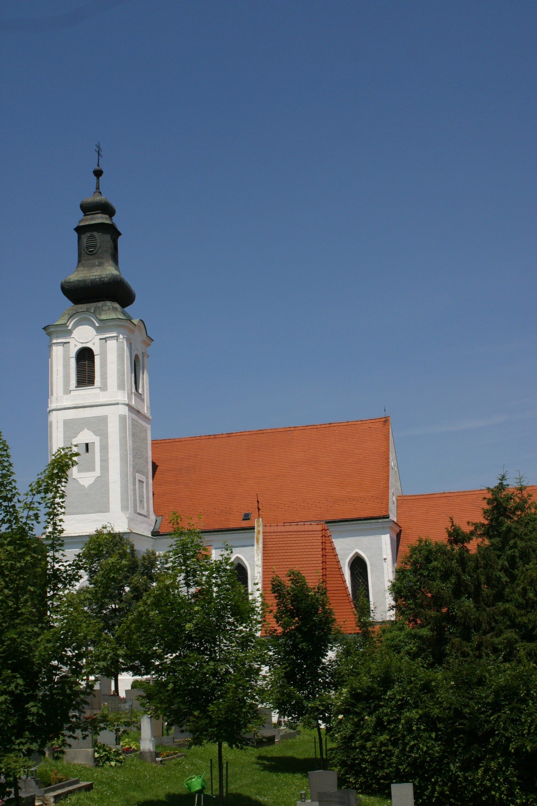 Eine Kirche mit einem weißen Turm und einem roten Dach ist unter einem klaren blauen Himmel zu sehen. Bäume umgeben das Gebäude.