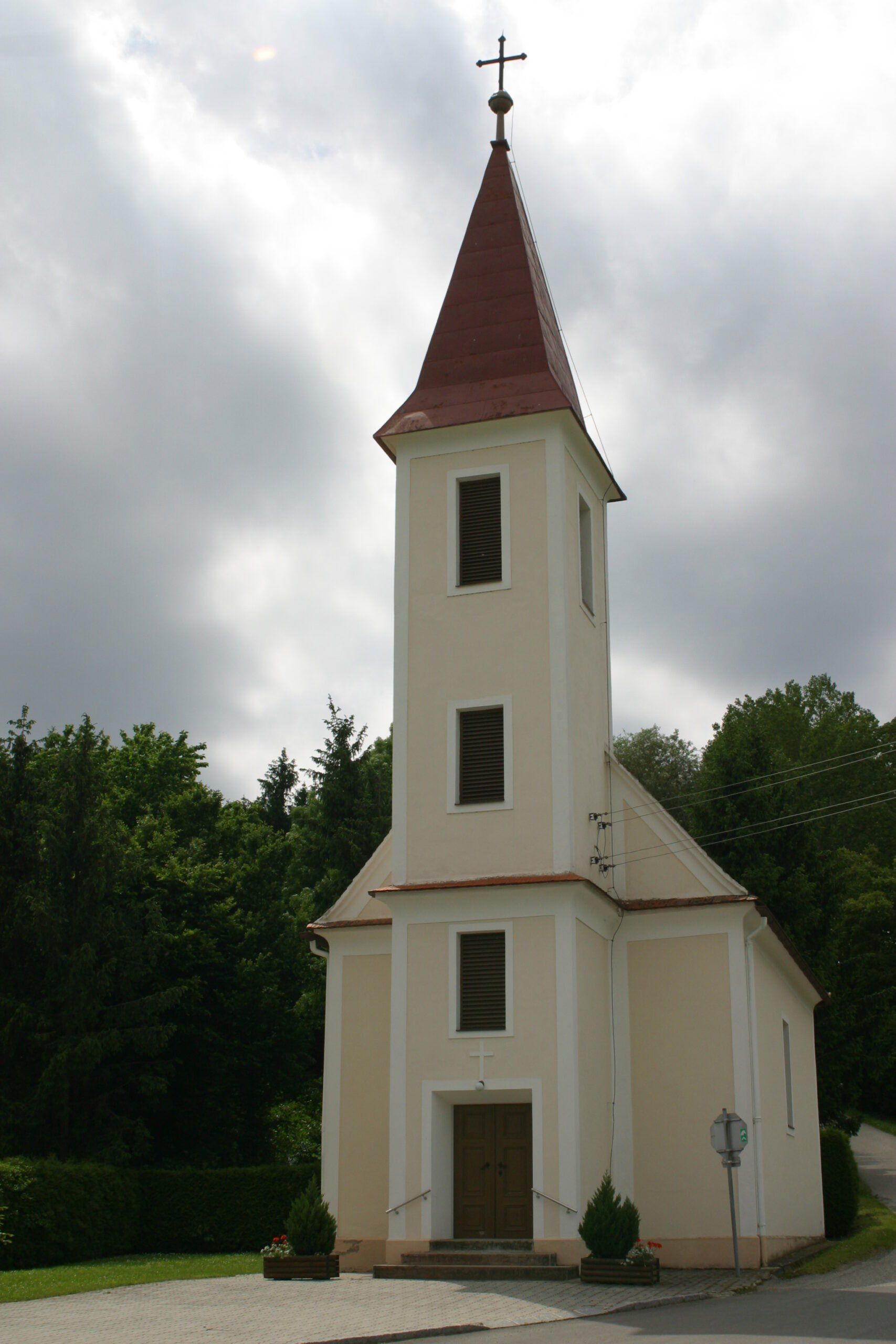 Eine kleine Kirche mit einem roten Dach und weißen Wänden steht inmitten grüner Bäume. Ein Kreuz befindet sich über der Tür, und es gibt Fenster auf dem Turm.