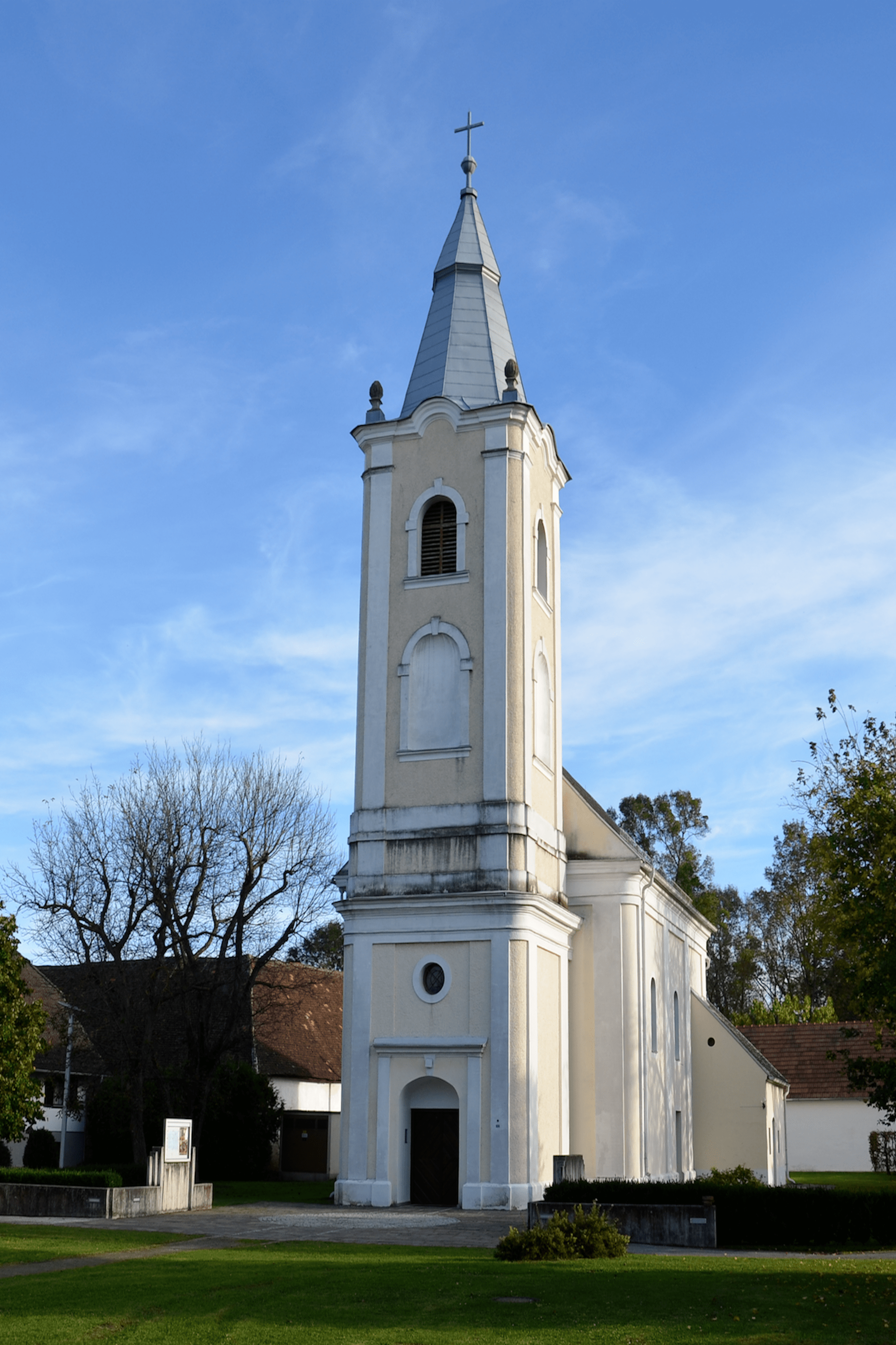 Ein hoher weißer Kirchturm steht unter einem klaren blauen Himmel. Der Turm hat mehrere Fenster und ein spitzes Dach. In der Nähe gibt es kahle Bäume und ein Haus mit einem braunen Dach.