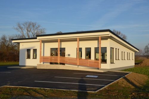 Ein modernes weißes Gebäude mit schwarzem Dach und großen Fenstern steht an einem sonnigen Tag. Das Gebäude hat Holzsäulen, die eine Rampe am Eingang stützen. Vor dem Gebäude befindet sich ein Parkplatz.