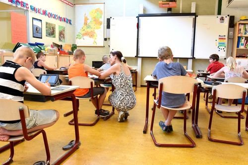 Ein Klassenzimmer mit Schülern an Tischen, einer Lehrerin, einer Tafel und Karten an der Wand.