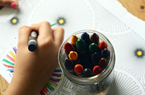 Eine Kinderhand hält einen Buntstift und färbt ein großes kreisförmiges Muster auf einem Papierblatt ein. Ein Glas mit Buntstiften ist mit verschiedenen Farben gefüllt und neben dem Papier platziert.