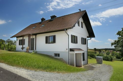 Ein weißes Haus mit braunem Dach und schwarzen Fensterläden befindet sich auf einem Hügel. Das Haus hat drei Fenster im Obergeschoss und drei im Erdgeschoss. Ein Fass ist rechts neben dem Haus platziert.