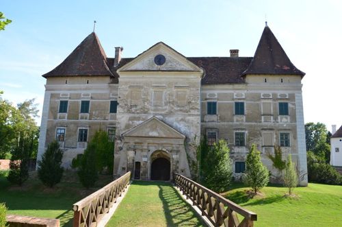 Ein altes Schloss mit braunen Dächern und vielen Fenstern, in einem grasbewachsenen Bereich mit einer Holzbrücke davor.