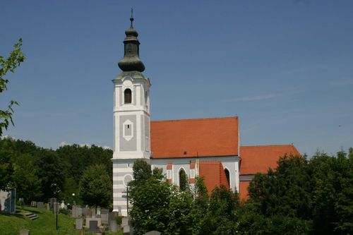 Eine Kirche mit Turm und rotem Dach steht gegen einen klaren blauen Himmel, umgeben von einem Friedhof und üppigen grünen Bäumen.