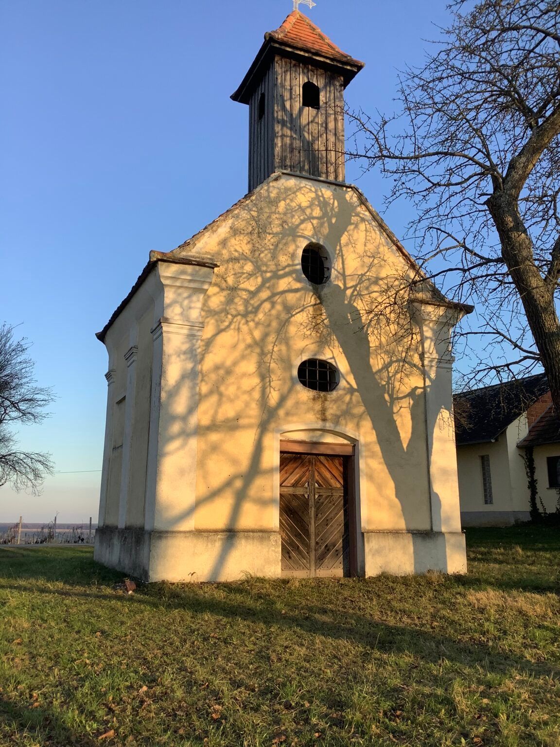 Eine kleine Kirche mit einer Holztür und Turm steht auf einem Rasenfeld. Schatten, die von Bäumen geworfen werden, sind auf dem Gebäude sichtbar.