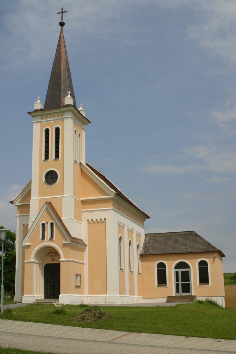 Eine beige Kirche mit einem spitzen Turm, Bogenfenstern und einem Schieferdach. Der Eingang hat eine Holztür mit einem Bogen darüber.