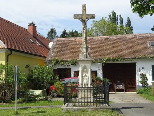 Ein Denkmal mit einem Kreuz und einer Statue steht vor einem Haus. Ein roter Wagen ist in der Nähe geparkt und ein Rasen umgibt das Denkmal.