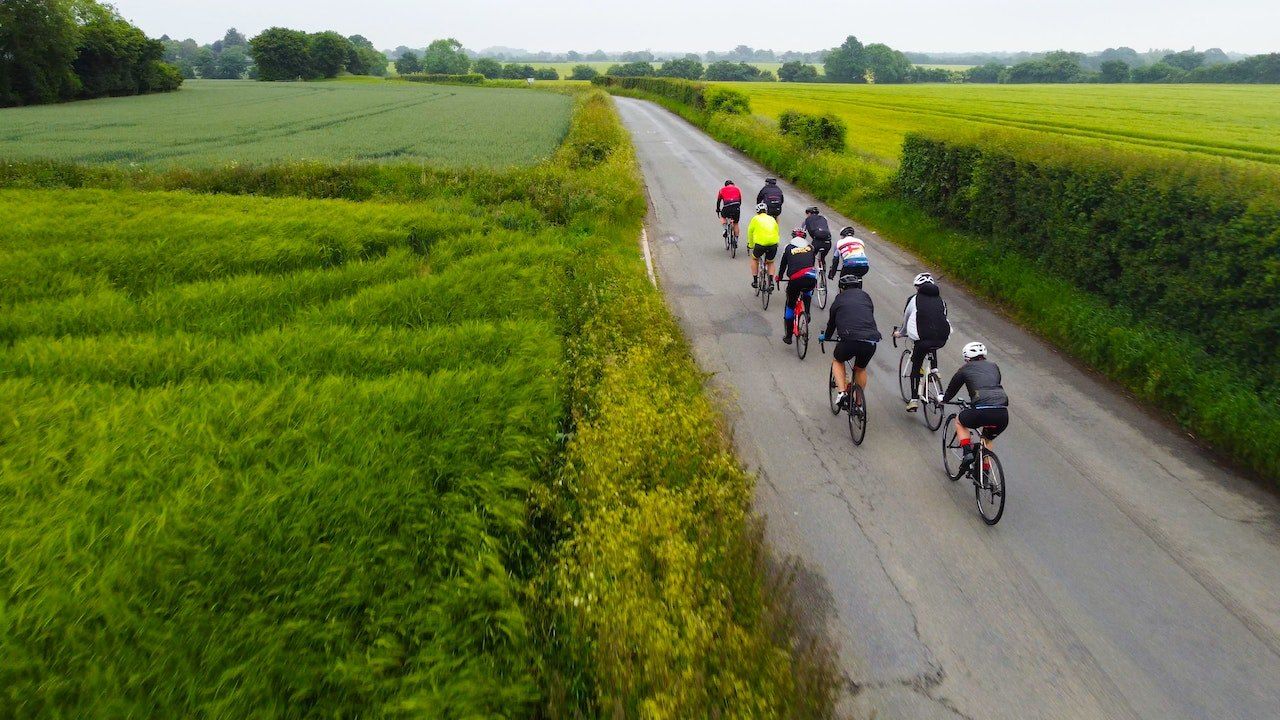 Eine Gruppe von Radfahrern fährt auf einer ländlichen Straße mit grünen Feldern auf beiden Seiten. Sie tragen Helme und Fahrradausrüstung.