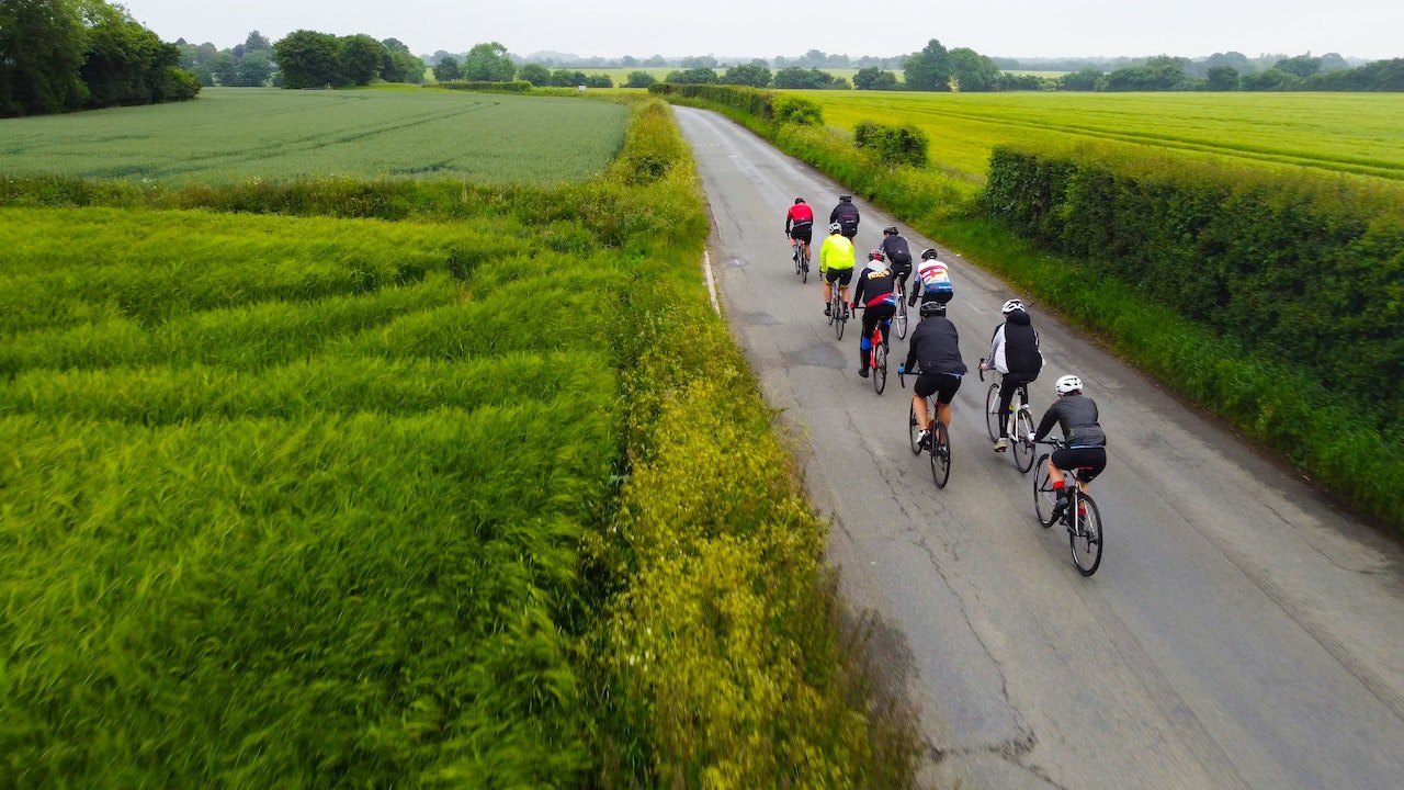 Eine Gruppe von Radfahrern fährt auf einer ländlichen Straße mit grünen Feldern auf beiden Seiten. Sie tragen Helme und Fahrradausrüstung.