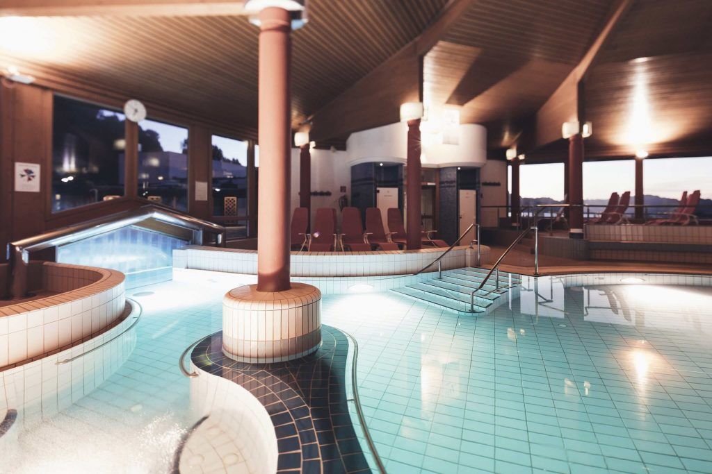 Ein Innenpool mit blauem Wasser, gefliestem Boden und Holzbalken. Rote Liegestühle und Stufen, die zum Pool führen. Eine Uhr und Lampen sind an der Wand montiert.
