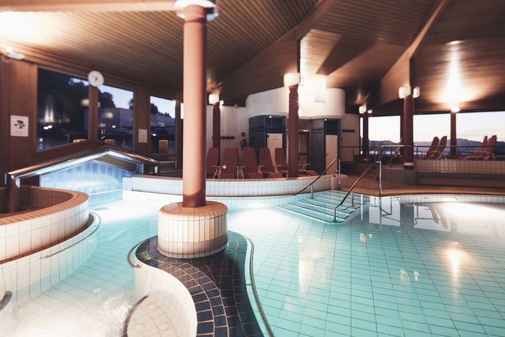 Ein Innenpool mit blauem Wasser, gefliestem Boden und Holzbalken. Rote Liegestühle und Stufen, die zum Pool führen. Eine Uhr und Lampen sind an der Wand montiert.