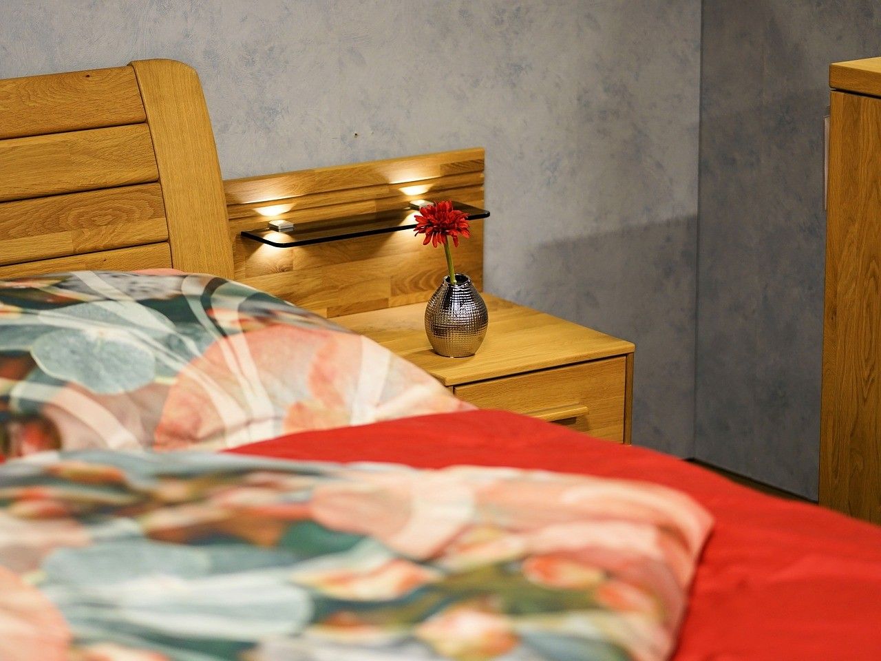 Ein Schlafzimmer mit einem Holzbett und Nachttisch. Der Nachttisch hat eine silberne Vase mit einer roten Blume. Das Kopfteil hat einen Holzrahmen mit LED-Leuchten.