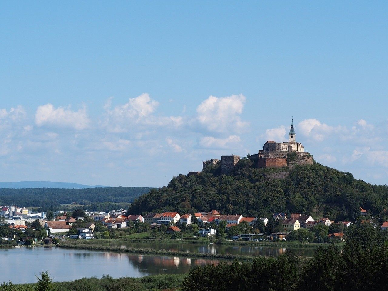 Eine Burg steht auf einem Hügel, umgeben von einer Stadt mit rot gedeckten Gebäuden, Bäumen und einem Fluss unter einem blauen Himmel mit Wolken.