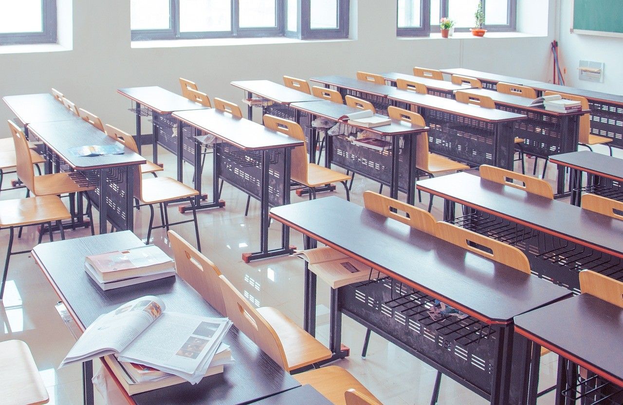 Ein leerer Klassenraum mit mehreren Tischen und Stühlen in Reihen angeordnet. Einige Bücher liegen auf den Tischen. Fenster mit Topfpflanzen an den Wänden.