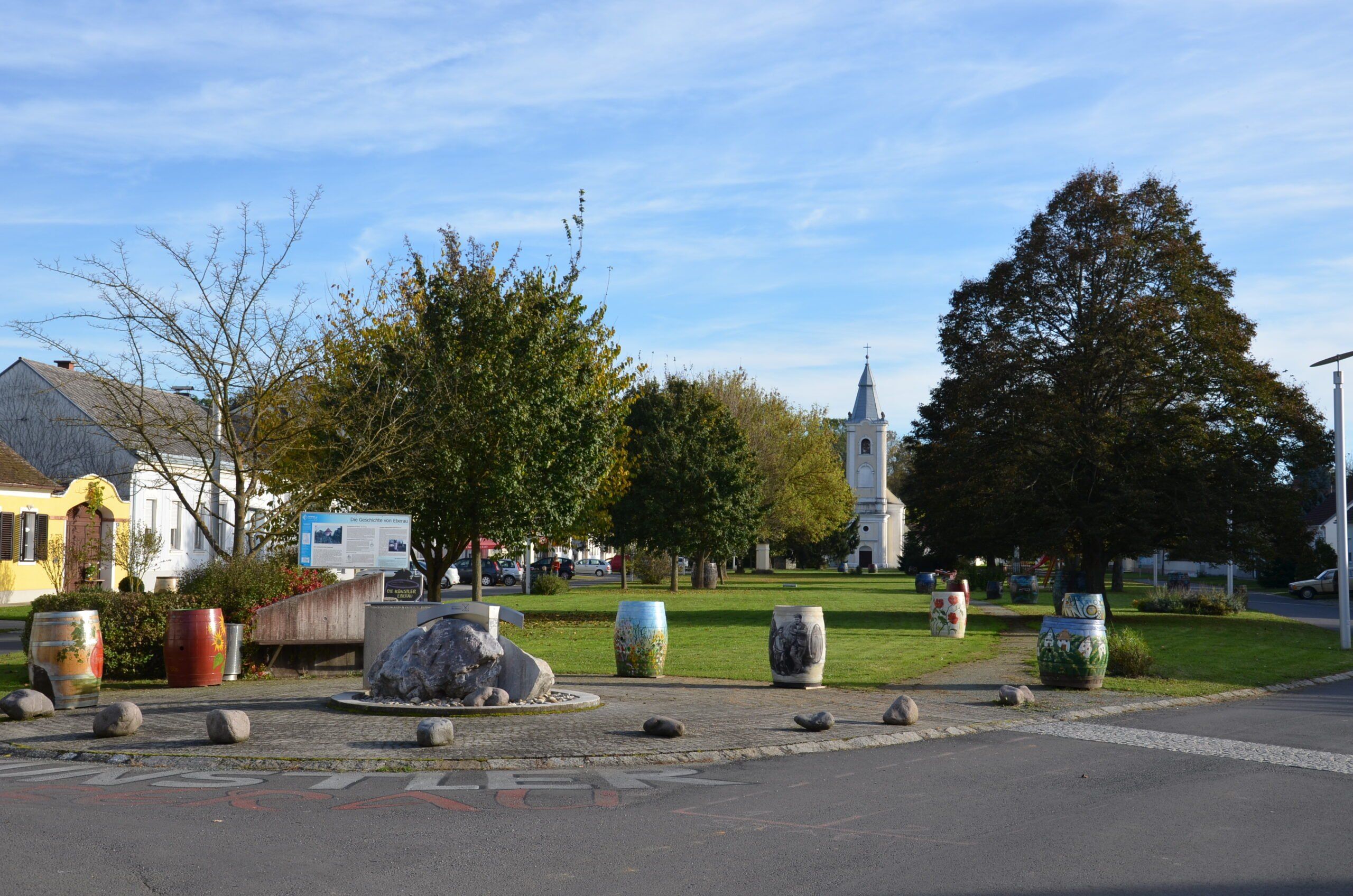 Ein Park mit Bäumen und einem Rasenbereich. Im Vordergrund befindet sich ein großer Felsen mit einer Tierskulptur. Am Straßenrand stehen bemalte Fässer. In der Ferne steht ein weißes Kirchengebäude.
