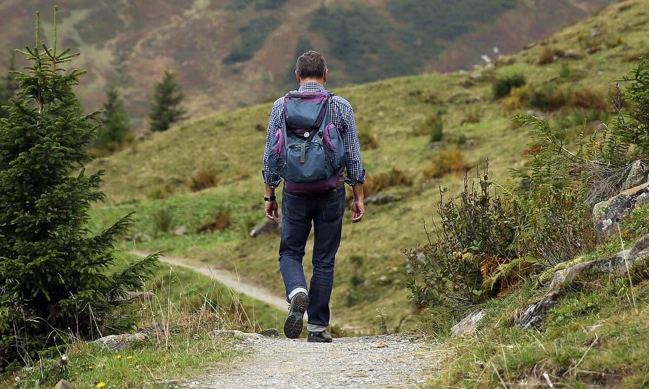Ein Mann mit Rucksack läuft auf einem Kiesweg in einem Grasfeld. Er schaut zurück und scheint in einem Gebirgsgebiet zu wandern.