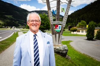 Ein älterer Mann mit Brille steht vor einem Holzschild mit den Worten 'Region Nockberge' und 'Patergassen'. Dahinter befinden sich Berge und ein klarer Himmel.