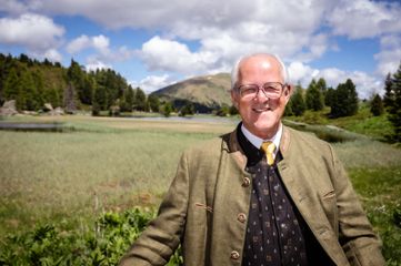 Ein älterer Mann mit Brille und gelber Krawatte steht in einem Grasfeld mit Bergen im Hintergrund.