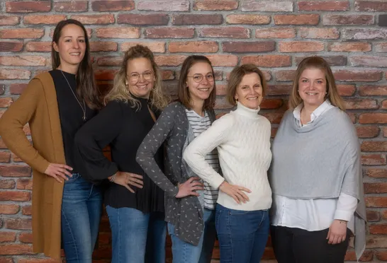 Fünf Frauen stehen zusammen vor einer Ziegelwand und lächeln für ein Foto. Sie tragen lässige Kleidung mit einer Mischung aus Oberteilen und Jeans. Die Frau in der Mitte trägt ein weißes Rollkragenshirt.