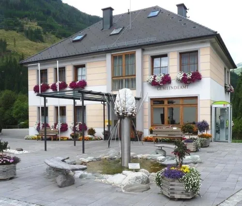 Ein Gebäude mit mehreren Fenstern und Balkonen, die mit Blumenkästen geschmückt sind, steht in der Nähe eines Brunnens, umgeben von Bänken und Topfpflanzen. Ein Schild lautet 'Gemeindeamt'.