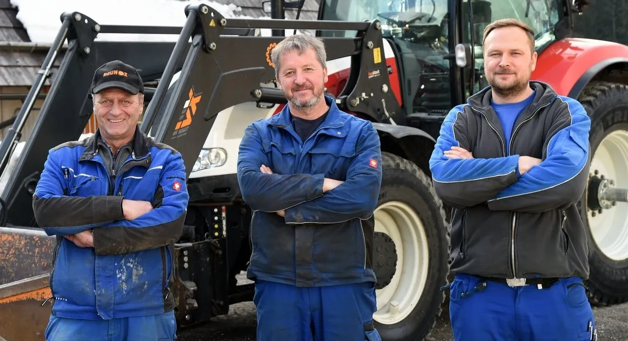 Drei Männer in blauer und schwarzer Arbeitskleidung stehen vor einem Traktor, lächeln und posieren für ein Foto. Der Mann in der Mitte hat die Arme verschränkt.