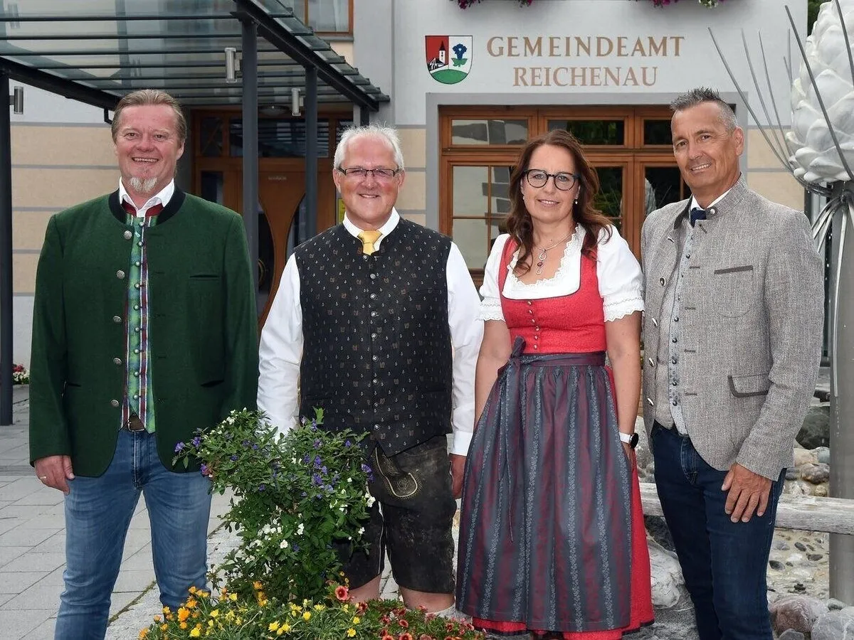 Vier Personen stehen vor dem Gemeindeamt Reichenau. Der Mann links trägt eine grüne Jacke. Der Mann in der Mitte trägt eine schwarze Weste. Die Frau trägt ein rotes und graues Kleid. Der Mann rechts trägt eine graue Jacke.