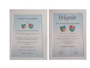Zwei gerahmte Dokumente, jedes mit dem Wort 'Urkunde' oben. Das linke Dokument zeigt den Text 'Ziele der Partnerschaft' und einige Logos. Das rechte Dokument hat Text in einer Fremdsprache, ein Datum und mehrere Unterschriften.