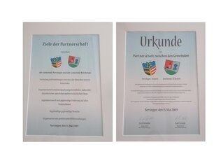 Zwei gerahmte Dokumente, jedes mit dem Wort 'Urkunde' oben. Das linke Dokument zeigt den Text 'Ziele der Partnerschaft' und einige Logos. Das rechte Dokument hat Text in einer Fremdsprache, ein Datum und mehrere Unterschriften.