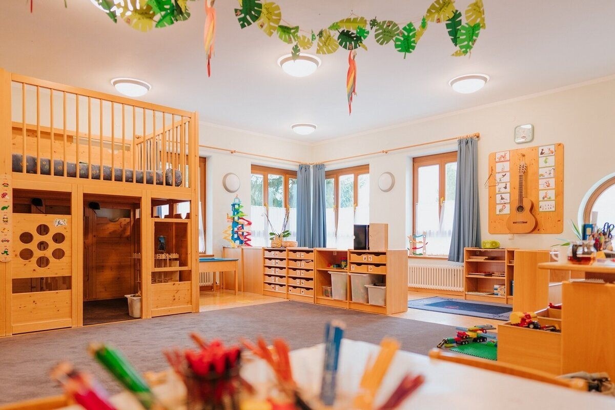 Ein Kinderzimmer mit einem Holzbett, Regalen und Spielzeug. Ein weißer Schreibtisch und Aufbewahrungsboxen in der Mitte, mit Pflanzen und einem Heizkörper in der Nähe. Blaue Vorhänge bedecken die Fenster, und ein blauer Teppich bedeckt den Boden.