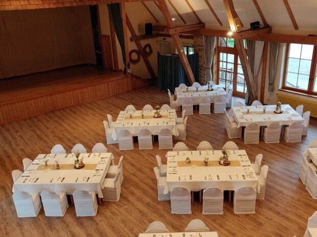 Hier sieht man den Saal des Nockstadls von oben. Man sieht einige Tische mit weißen Tüchern gedeckt und Stühle daneben. Die Tische sich mit Besteck und Teller gedeckt und mit Blumen