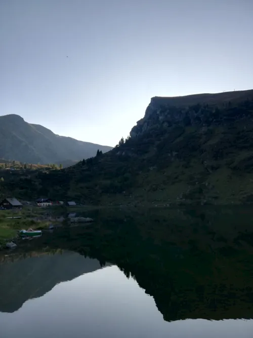 Ein ruhiger Bergsee mit Spiegelungen der umliegenden Berge und einigen Gebäuden. Der Himmel ist klar und blau.