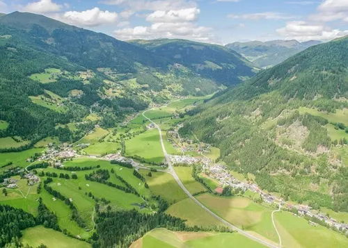 Eine landschaftliche Luftaufnahme eines Tals mit grünen Wiesen, einer kurvenreichen Straße und Gebäudeclustern, umgeben von üppigen Wäldern und Bergen unter einem teilweise bewölkten Himmel.