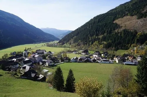 Luftaufnahme eines kleinen Dorfes in einem Tal, umgeben von saftig grünen Feldern und Kiefernwäldern unter einem klaren blauen Himmel.