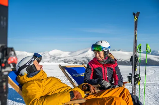 Zwei Personen in Skiausrüstung sitzen auf Liegestühlen im Schnee, mit einem blauen Stuhl dahinter und verschneiten Bergen im Hintergrund.