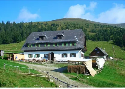 Ein Haus mit grauem Dach und grünen Fensterläden steht in der Mitte eines grünen Feldes. Das Feld ist von Bäumen umgeben.