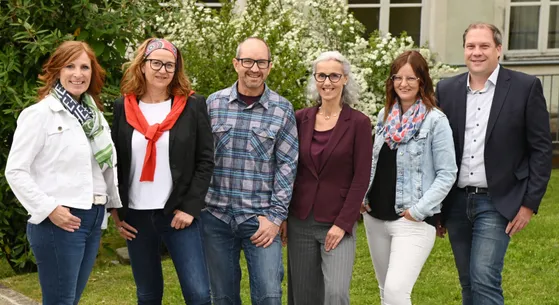 Vier Erwachsene, zwei Männer und zwei Frauen, lächeln und posieren für ein Foto in einem Garten mit weißen Blumen im Hintergrund.