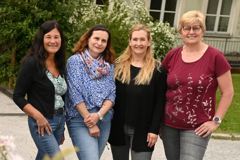 Fünf Frauen stehen zusammen draußen. Sie lächeln und scheinen für ein Foto zu posieren. Hinter ihnen befinden sich Pflanzen und ein Gebäude.
