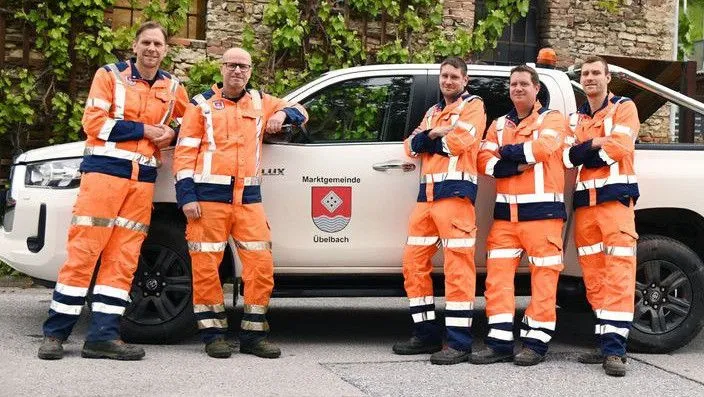 Fünf Männer in orangefarbenen Uniformen stehen vor einem weißen Pickup-Truck mit der Aufschrift 'Marktgemeinde Uttelbach'. Sie posieren für ein Foto.