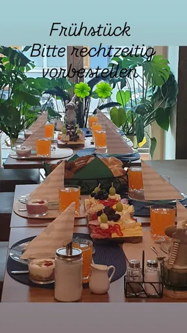Ein langer Tisch ist mit Brot, Käse, Oliven und Getränken gedeckt. Im Hintergrund sind grüne Pflanzen und ein vierblättriges Kleeblatt zu sehen.