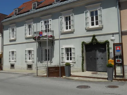 Ein hellblaues Gebäude mit weißen Fensterläden und einem Balkon mit einem roten Schild. Eine graue Tür mit einem Kranz und zwei Topfpflanzen stehen davor.