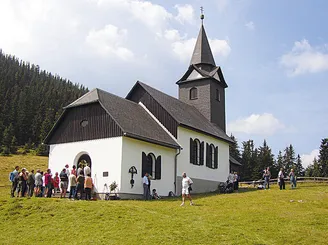 Eine Kirche mit einem Turm steht auf einem Hügel, umgeben von saftigem grünem Gras und Bäumen. Mehrere Menschen versammeln sich in der Nähe des Eingangs, während andere gehen oder stehen. Der Himmel ist teilweise bewölkt.