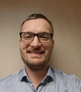 Ein Mann mit Brille, Bart und kurzem Haarschnitt, lächelt und posiert für ein Foto. Er trägt ein gemustertes Hemd und hat eine helle Wand hinter sich.