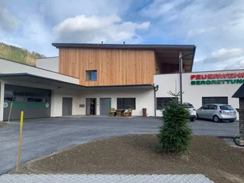 Die Vorderseite eines modernen Gebäudes mit Holzfassade. Das Schild sagt Feuer Berg. Eine Person steht am Eingang, und zwei Autos sind davor geparkt.