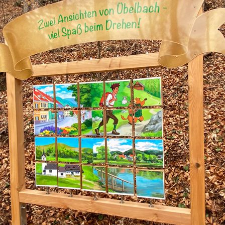 Ein Holzbrett mit verschiedenen Illustrationen darauf, umgeben von einem braunen Band. Das Band trägt die Aufschrift 'Zwei Ansichten von ?belbach - viel Spa? beim Drehen!'. Die Bilder zeigen einen Wald, einen Fluss, ein Haus, eine Person, die geht, und eine Brücke.